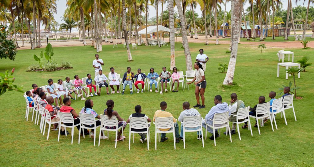 Les enfants assis en cercle sur la pelouse, sous les cocotiers, au camp Mi-Adja 2025 à San Pedro