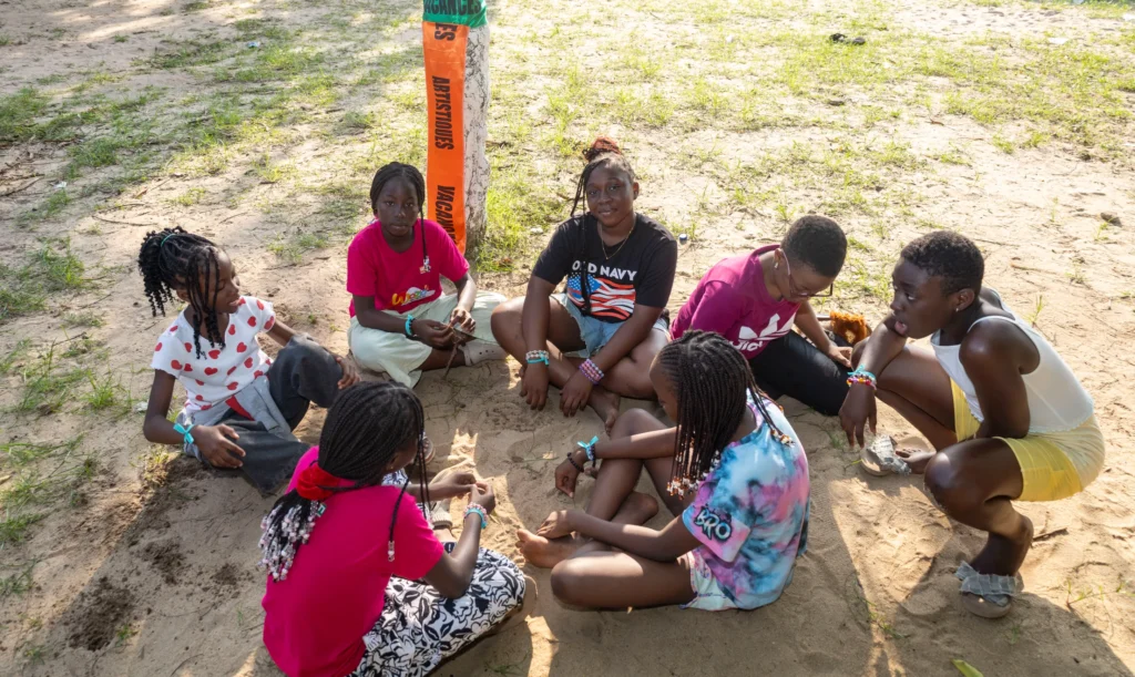 Des enfants assis en cercle sur le sable discutent, au camp Mi-Adja 2025 à San Pedro